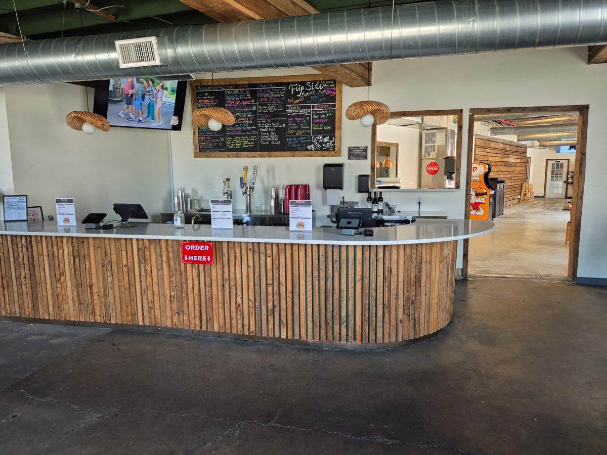 The order counter at Flip Side Burger Bar with the hand-lettered chalkboard menu above it, draft taps, and the arcade visible down the hall
