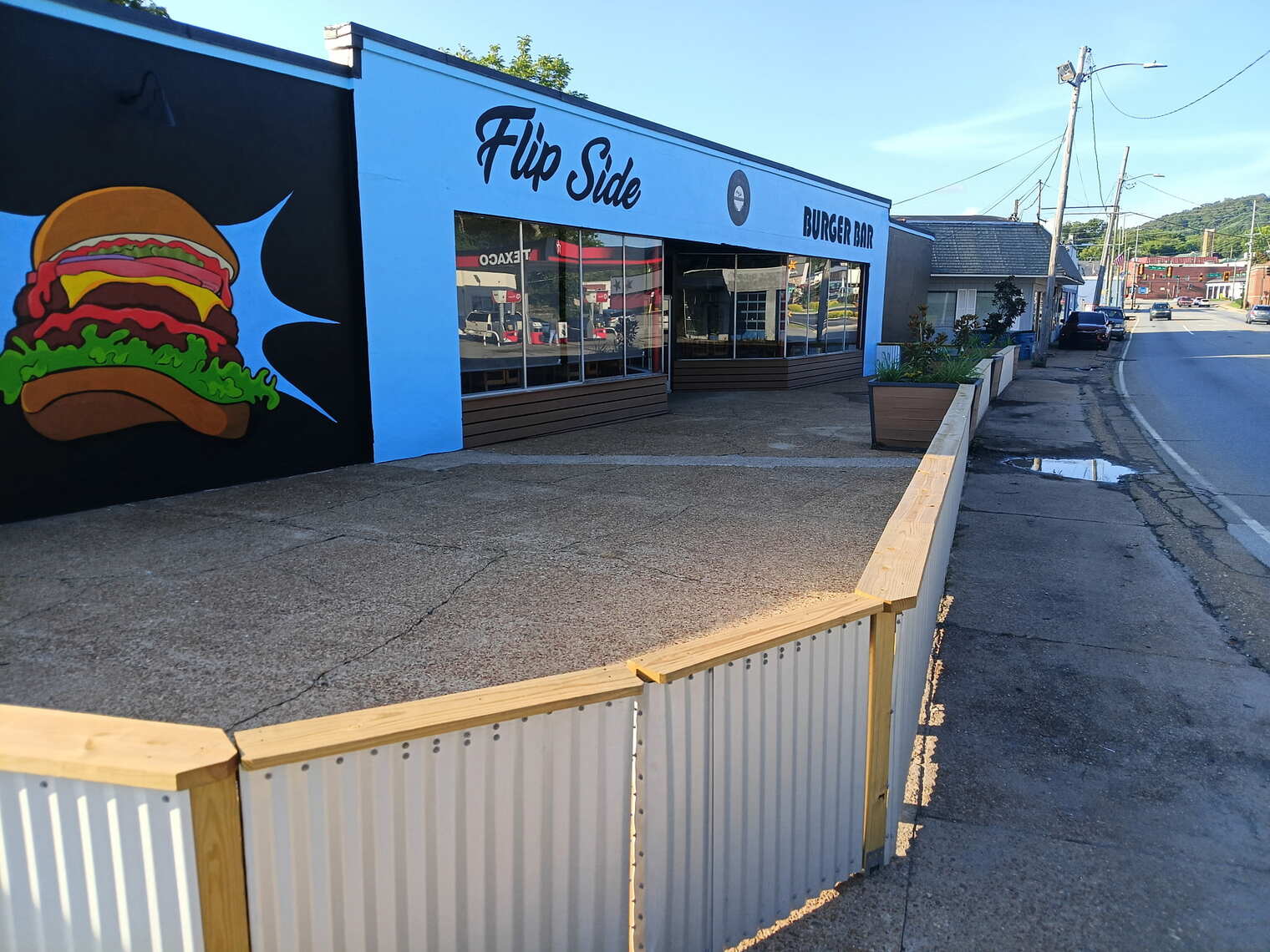 Flip Side Burger Bar's storefront during the day — a bright blue building with signage, the burger mural on the black side wall, and patio fence