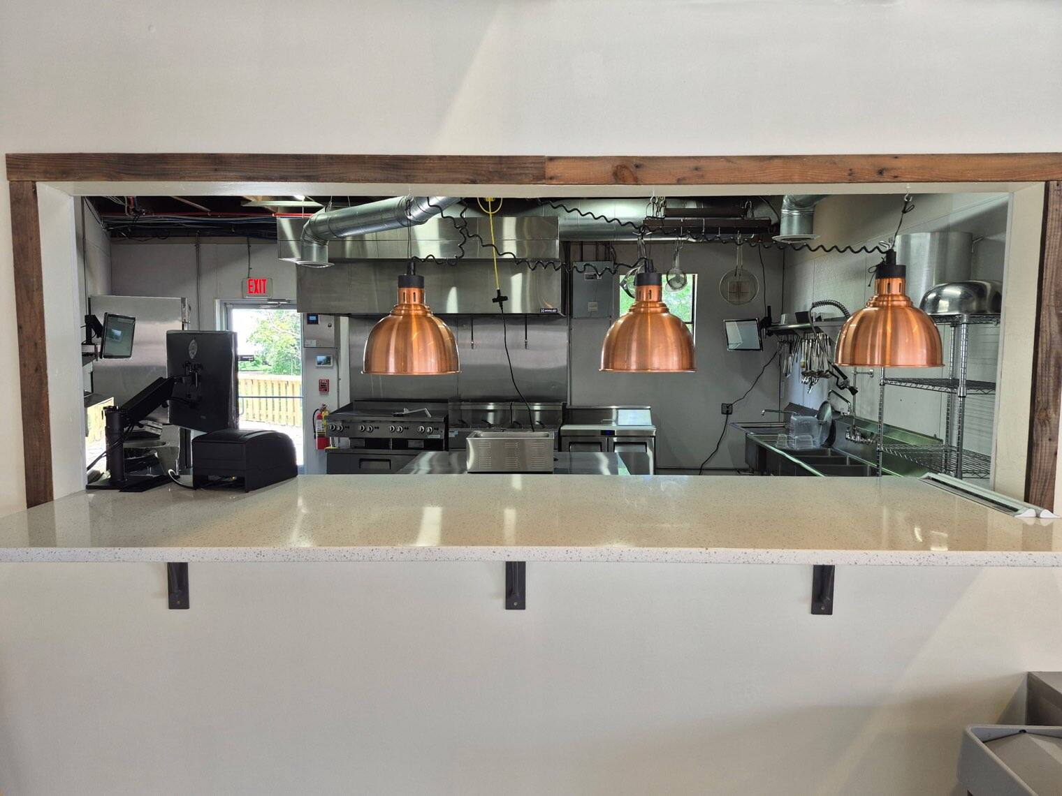 The kitchen pass at Flip Side Burger Bar &mdash; three copper heat lamps over a white quartz counter looking into the stainless-steel kitchen
