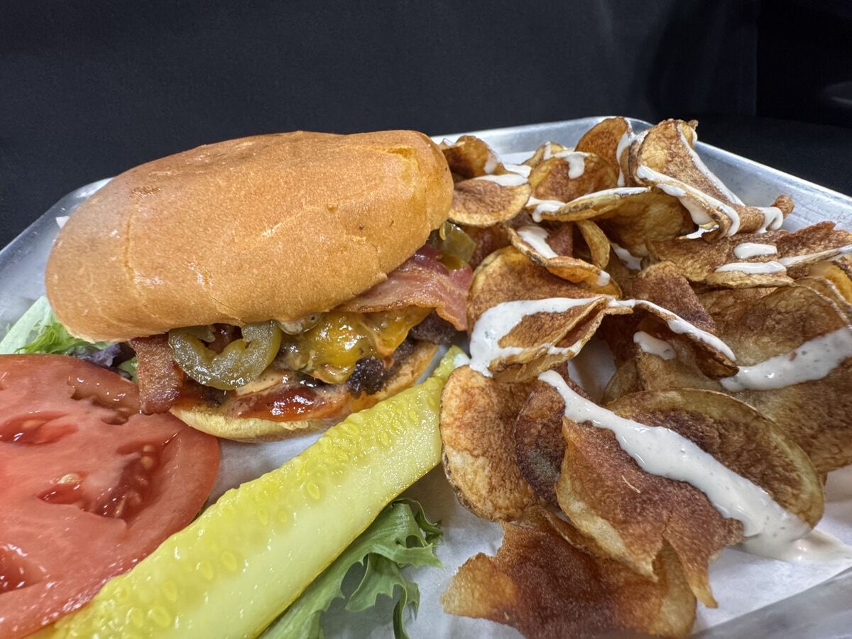The Cowboy &mdash; smash patty with melted pepper jack, bacon, and candied jalape&ntilde;os in house BBQ on a brioche bun, served on a metal tray with ranch-drizzled house chips, a pickle spear, and tomato