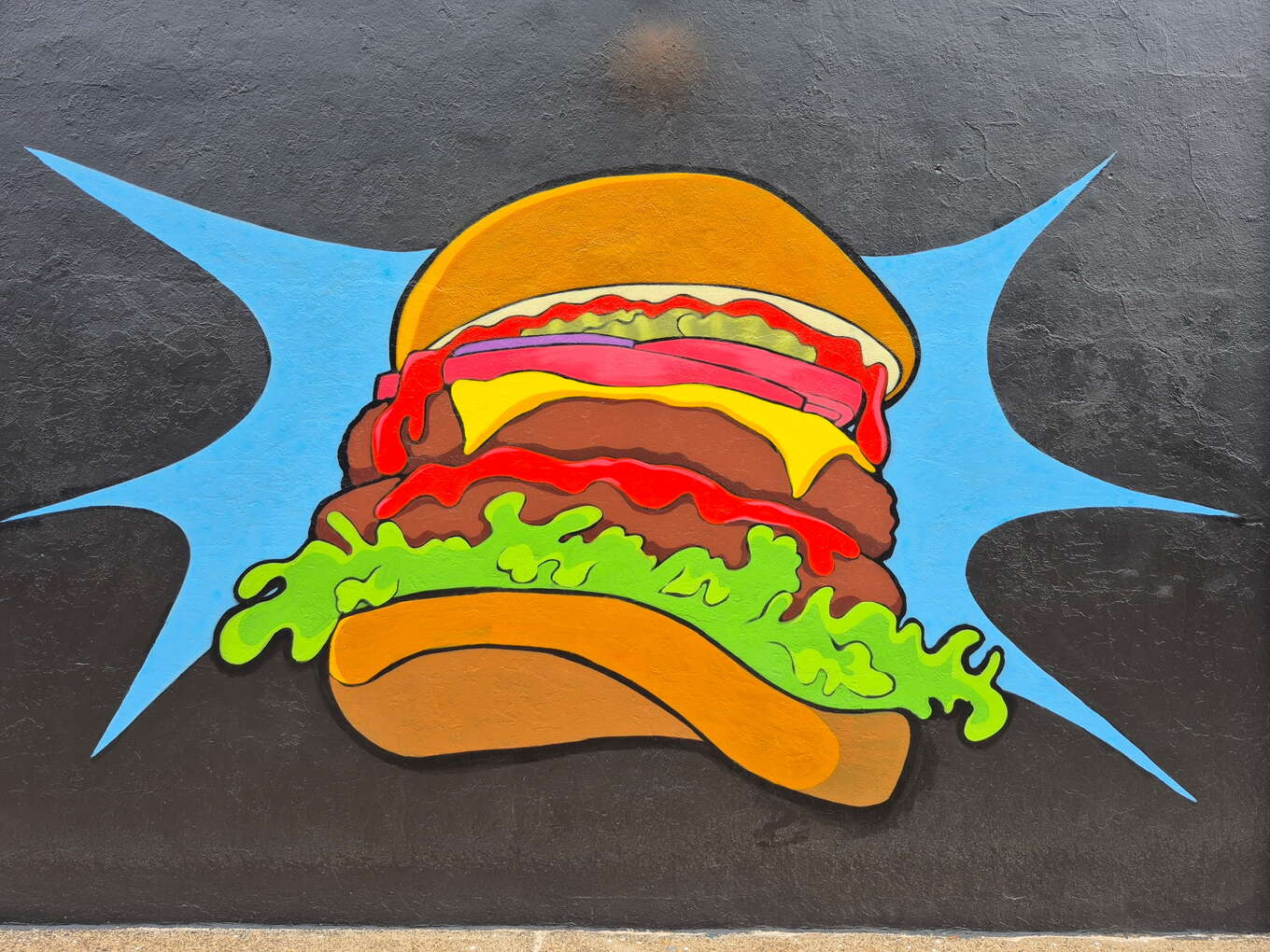 Close-up of the hand-painted cartoon cheeseburger mural on Flip Side Burger Bar's exterior wall, done in bold black outline with a blue starburst